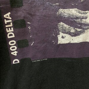 Pu&ograve; includere: Una maglietta nera con un'immagine viola di un uomo che suona la chitarra. Il testo "D 400 DELTA" &egrave; stampato sul lato sinistro della maglietta. Il testo "&copy; 1992 ERIC CLAPTON" &egrave; stampato sul lato inferiore destro della maglietta.