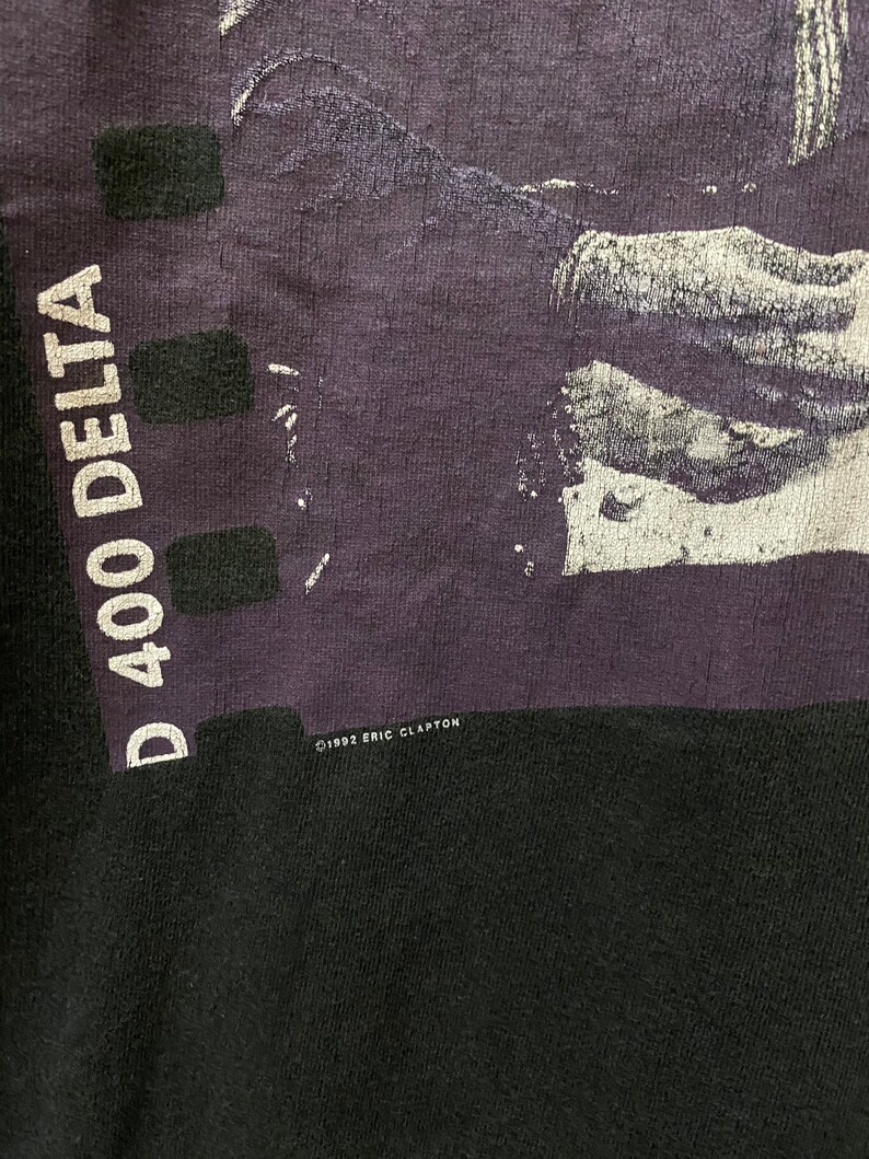 Pu&ograve; includere: Una maglietta nera con un'immagine viola di un uomo che suona la chitarra. Il testo "D 400 DELTA" &egrave; stampato sul lato sinistro della maglietta. Il testo "&copy; 1992 ERIC CLAPTON" &egrave; stampato sul lato inferiore destro della maglietta.