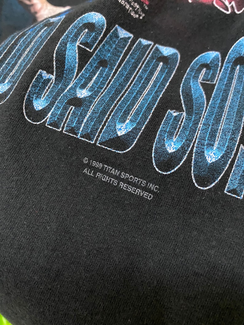 May include: Black t-shirt with a blue and white graphic that says "I Saw" with the text "&copy; 1998 TITAN SPORTS INC. ALL RIGHTS RESERVED" below.