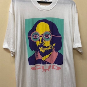 May include: White t-shirt featuring a colourful graphic print of a man wearing glasses with large eyes. The text "CUD" is printed below the graphic.