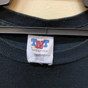 May include: A black t-shirt tag with white text that reads "TNT", "AU & IQEVNER", "ADULT - L", "100% COTTON", "MADE IN MEXICO", and "CARE ON REVERSE".
