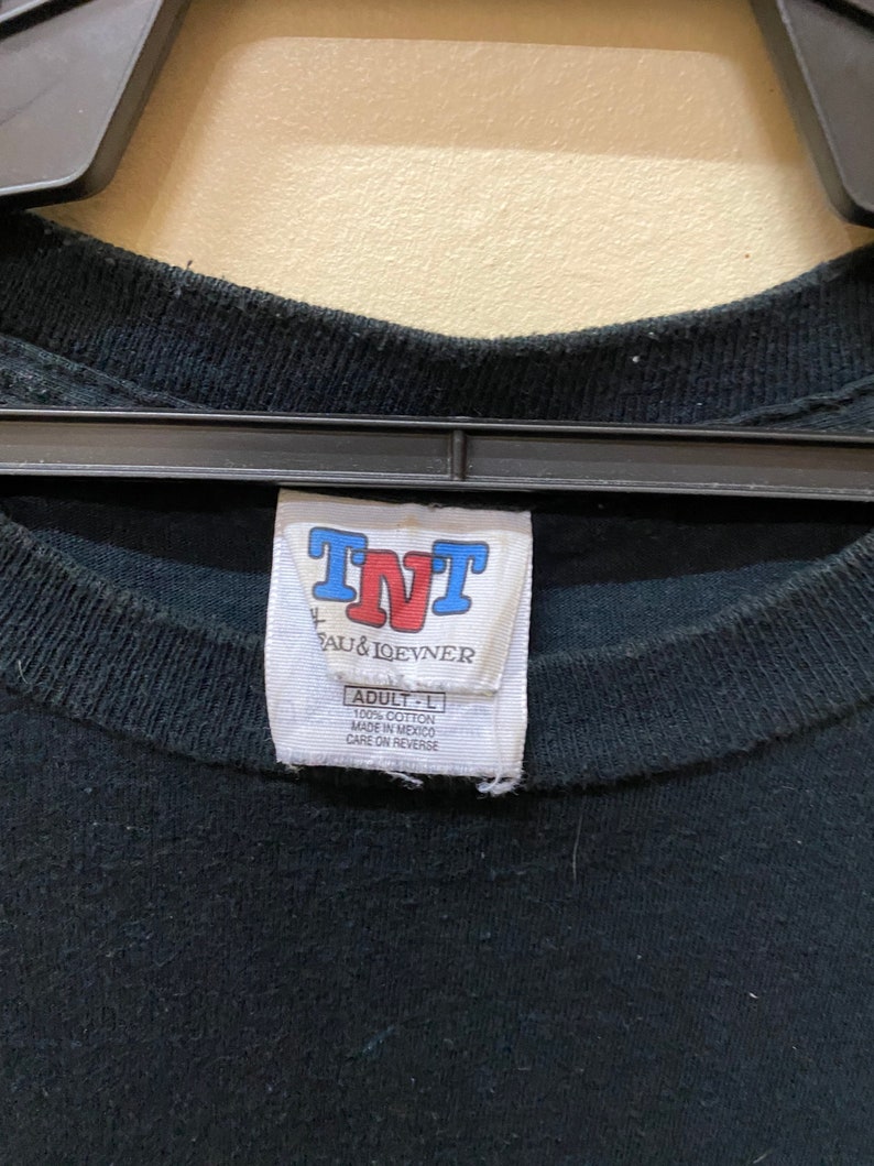 May include: A black t-shirt tag with white text that reads "TNT", "AU & IQEVNER", "ADULT - L", "100% COTTON", "MADE IN MEXICO", and "CARE ON REVERSE".