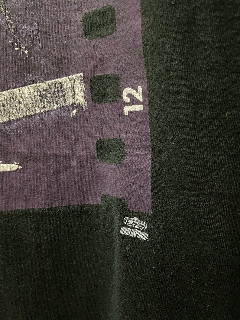 Pu&ograve; includere: Maglietta nera con una grafica viola e bianca di una pellicola con il numero 12 stampato su di essa. La grafica include anche il testo "Nintendo" e "High-Impact".