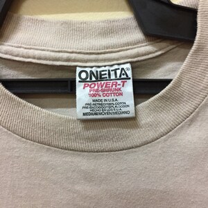 Puede incluir: Una camiseta beige con un estampado gr&aacute;fico en blanco y negro de una persona sosteniendo una c&aacute;mara. La camiseta tiene una etiqueta que dice "ONEITA POWER-T PRE-SHRUNK 100% COTTON MADE IN U.S.A. PRE-R&Eacute;TR&Eacute;CI/100% COTON PRE-ENCOGIDO/100% ALGODON HECHO EN LOS EUA. MEDIUM/MOYEN/MEDIANO"