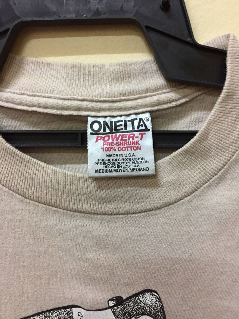 Puede incluir: Una camiseta beige con un estampado gr&aacute;fico en blanco y negro de una persona sosteniendo una c&aacute;mara. La camiseta tiene una etiqueta que dice "ONEITA POWER-T PRE-SHRUNK 100% COTTON MADE IN U.S.A. PRE-R&Eacute;TR&Eacute;CI/100% COTON PRE-ENCOGIDO/100% ALGODON HECHO EN LOS EUA. MEDIUM/MOYEN/MEDIANO"