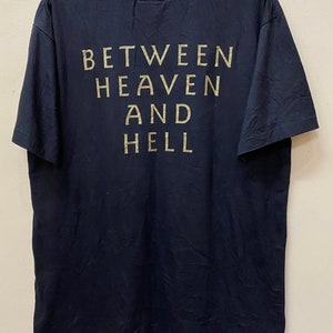 Puede incluir: Camiseta negra con el texto "BETWEEN HEAVEN AND HELL" impreso en dorado en la parte posterior.