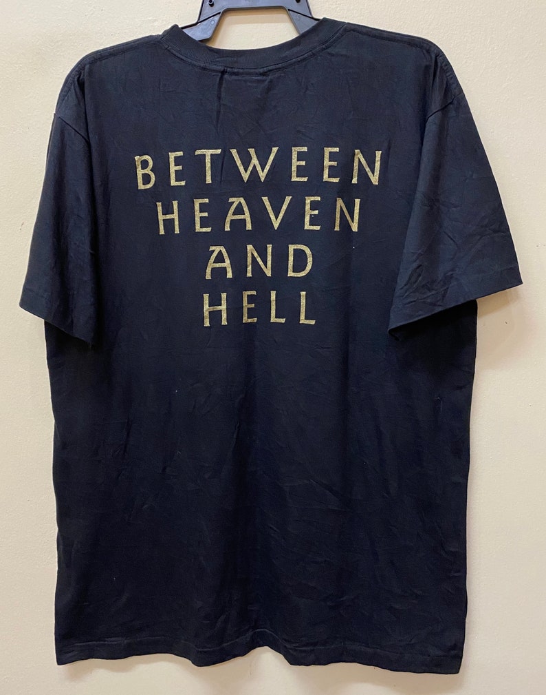 Puede incluir: Camiseta negra con el texto "BETWEEN HEAVEN AND HELL" impreso en dorado en la parte posterior.