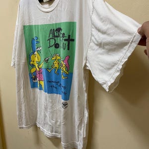 Puede incluir: Camiseta blanca con un gr&aacute;fico verde, azul y rosa de personajes de dibujos animados. El texto "Alice Donut" est&aacute; impreso en negro en el gr&aacute;fico. El texto "revenge of the flies" est&aacute; impreso en negro debajo del gr&aacute;fico.