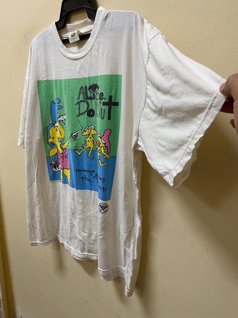 Puede incluir: Camiseta blanca con un gr&aacute;fico verde, azul y rosa de personajes de dibujos animados. El texto "Alice Donut" est&aacute; impreso en negro en el gr&aacute;fico. El texto "revenge of the flies" est&aacute; impreso en negro debajo del gr&aacute;fico.