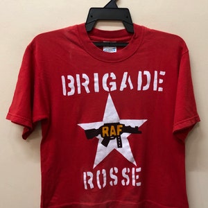 Pu&ograve; includere: Maglietta rossa con una grafica bianca di una stella con una pistola e il testo "BRIGADE ROSSE" e "RAF" al centro.
