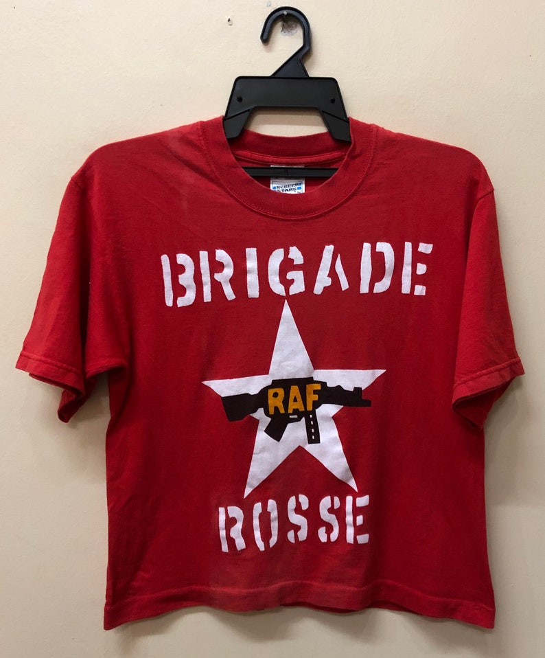 Pu&ograve; includere: Maglietta rossa con una grafica bianca di una stella con una pistola e il testo "BRIGADE ROSSE" e "RAF" al centro.