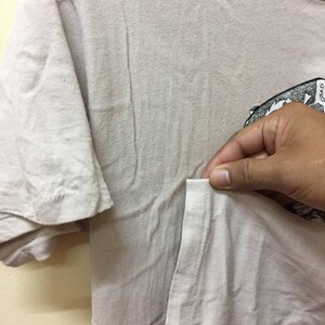 Puede incluir: Una camiseta beige claro con un estampado gr&aacute;fico en blanco y negro en el lado derecho. La camiseta tiene un bolsillo en el lado izquierdo.