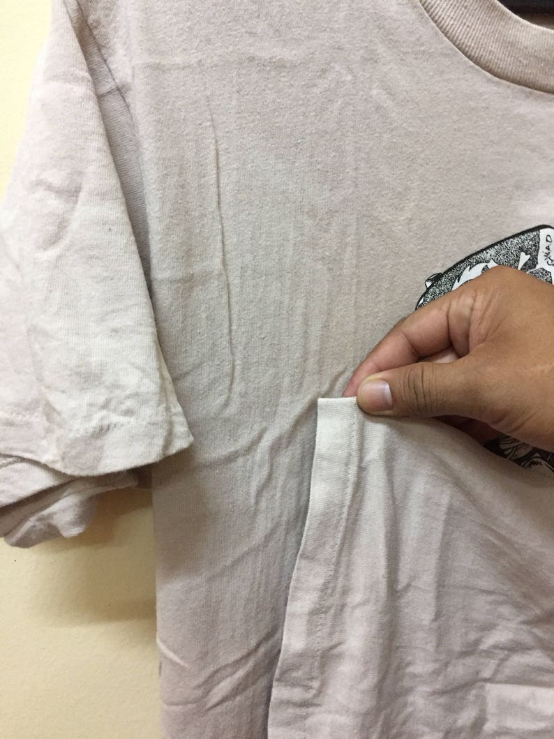 Puede incluir: Una camiseta beige claro con un estampado gr&aacute;fico en blanco y negro en el lado derecho. La camiseta tiene un bolsillo en el lado izquierdo.