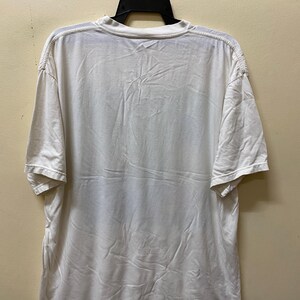 Puede incluir: Camiseta blanca de manga corta colgada de una percha negra. La camiseta es lisa, sin gr&aacute;ficos ni texto visibles.