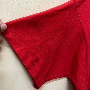 May include: Red short-sleeved t-shirt with a faded, worn look. The shirt has a few stains on the sleeve.