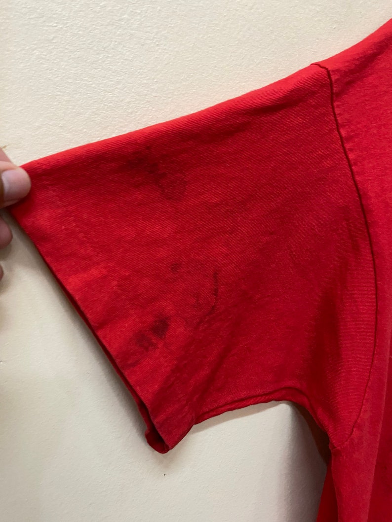 May include: Red short-sleeved t-shirt with a faded, worn look. The shirt has a few stains on the sleeve.