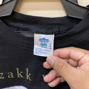 Puede incluir: Camiseta negra con una impresi&oacute;n dorada del texto "zakk wylde" y una imagen en blanco y negro de una calavera. La etiqueta de la camiseta dice "Winterland Productions Worldwide" y "Pre-shrunk 100% Cotton Made in USA".