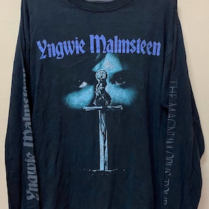 May include: Black long-sleeved t-shirt with a blue and white image of a sword and a face with the text "Yngwie Malmsteen" and "The Magnum Opus" on the front and sleeves.