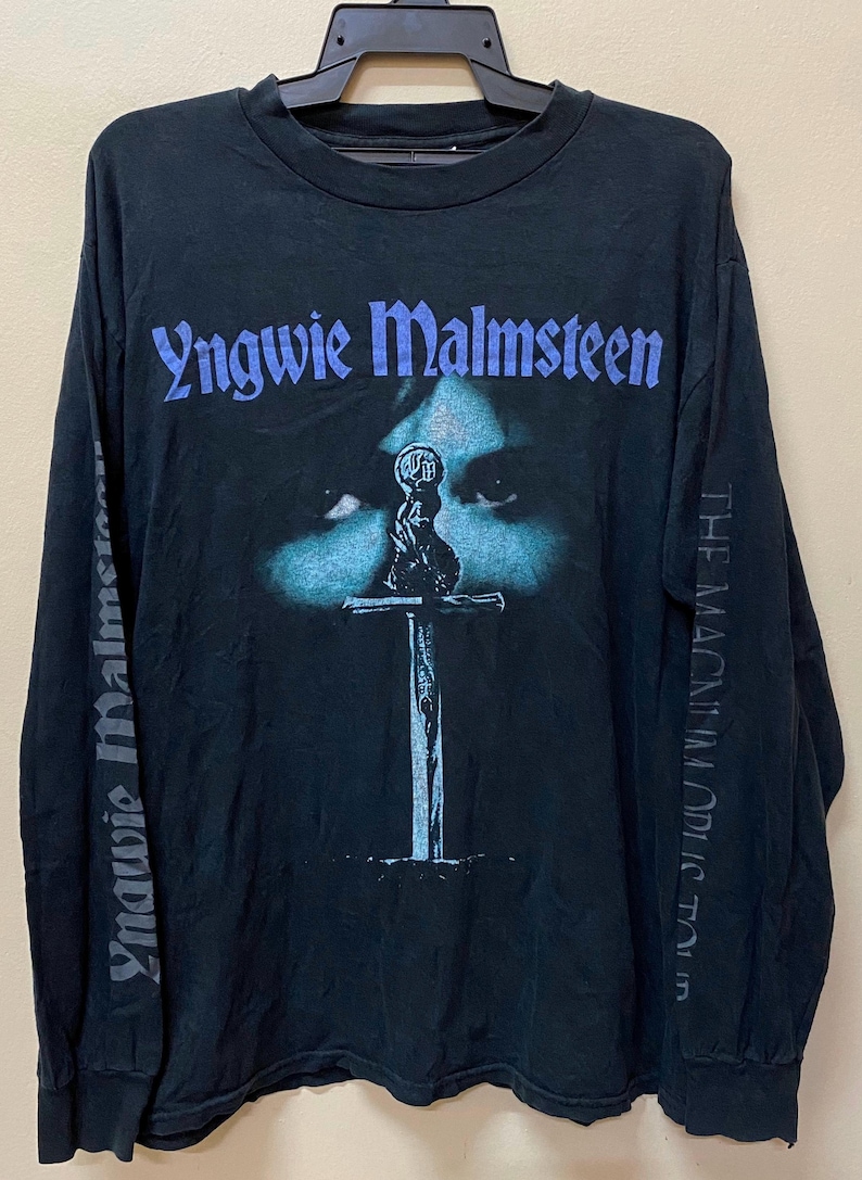 May include: Black long-sleeved t-shirt with a blue and white image of a sword and a face with the text "Yngwie Malmsteen" and "The Magnum Opus" on the front and sleeves.