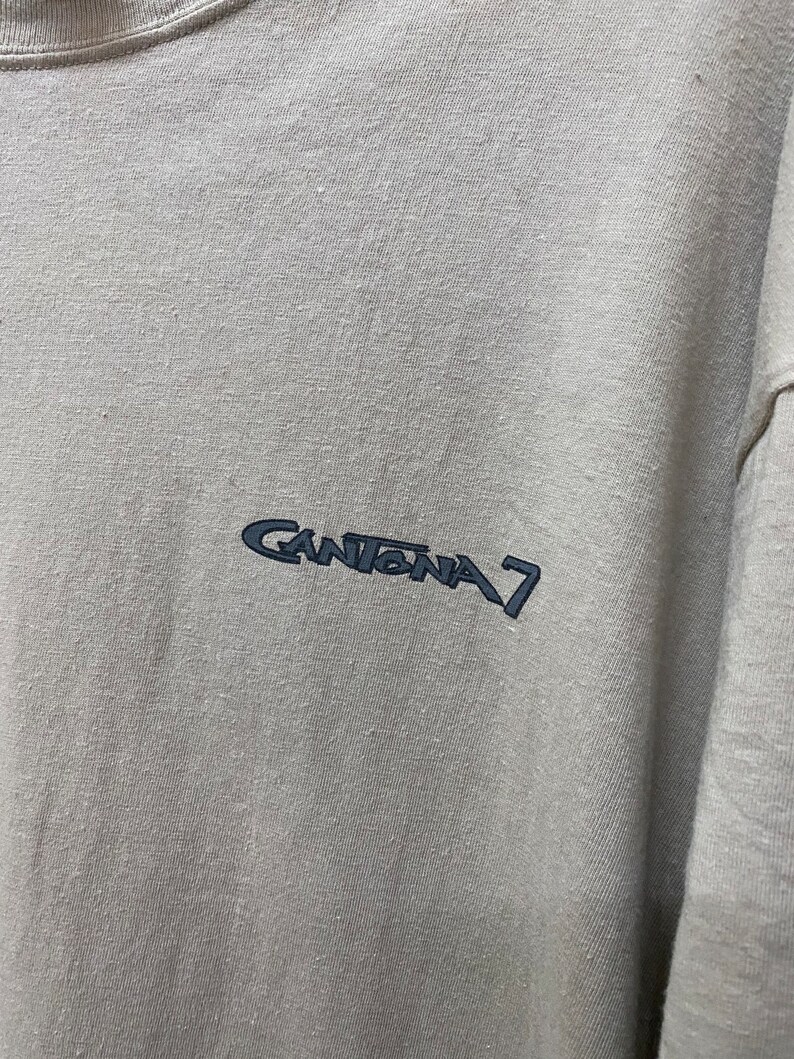 Może przedstawiać: Jasnoszary t-shirt z haftowanym czarnym logo z napisem "Cantona 7".