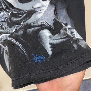 May include: Black t-shirt with a graphic print of a person's face. The print is in shades of gray and white. The t-shirt has a blue signature that says "Viagra 95".