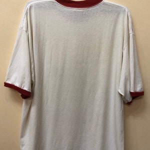 Puede incluir: Camiseta blanca con cuello y mangas de color rojo. La camiseta est&aacute; colgada en una percha negra.