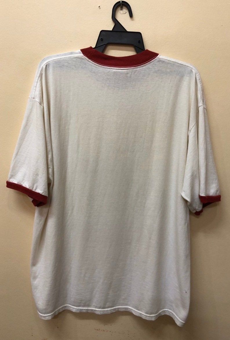 Puede incluir: Camiseta blanca con cuello y mangas de color rojo. La camiseta est&aacute; colgada en una percha negra.