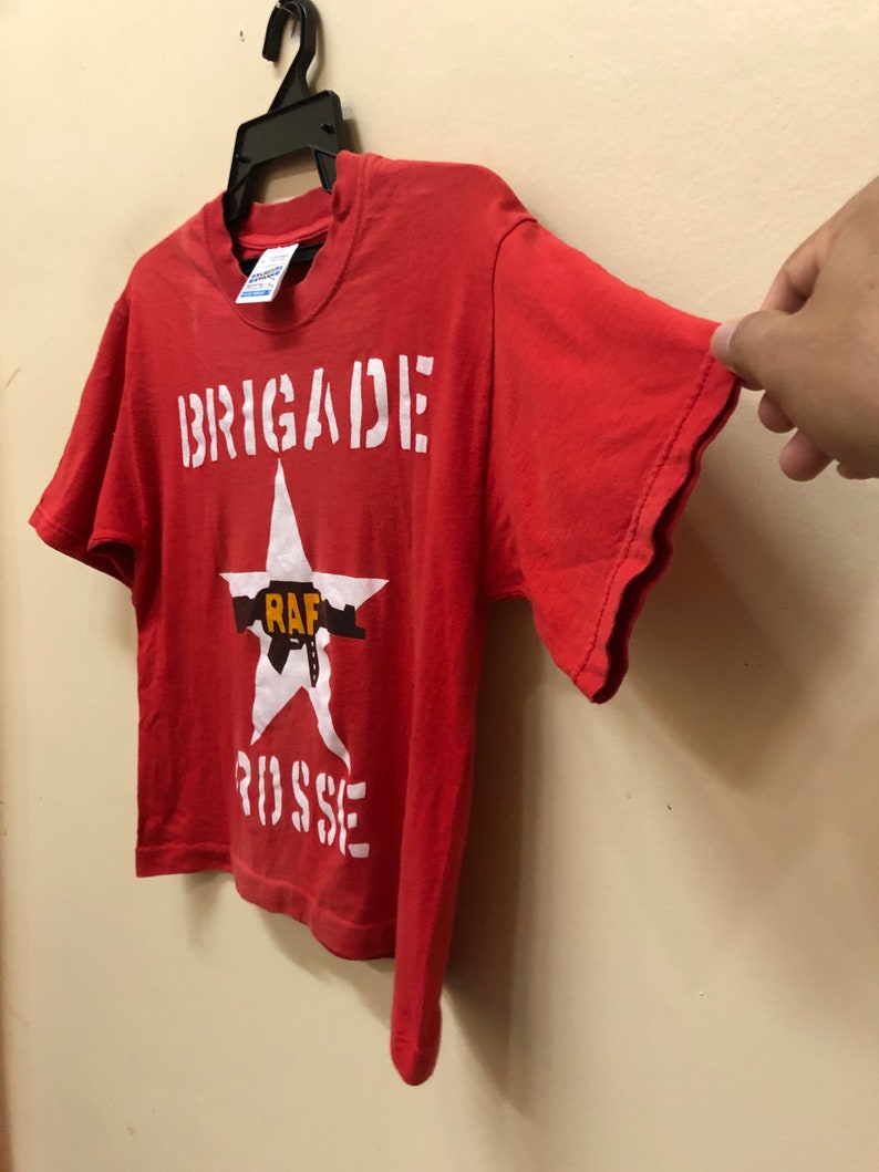 Pu&ograve; includere: Maglietta rossa a maniche corte con una stella bianca e il testo "BRIGADE RAF RUSSE" stampato sul davanti.
