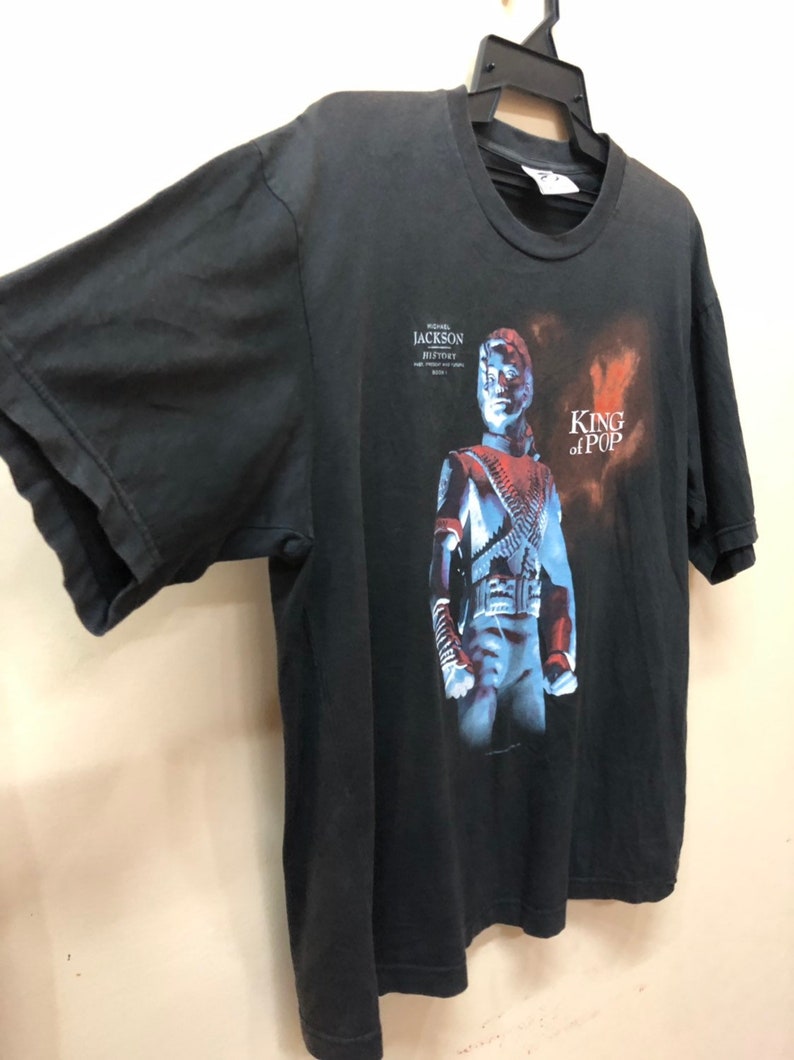 May include: Black t-shirt with a graphic print of Michael Jackson from his "History" album. The text "King of Pop" is printed on the shirt.