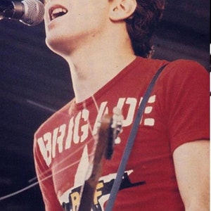 Pu&ograve; includere: Un uomo con i capelli corti e castani canta in un microfono mentre tiene una chitarra. Indossa una maglietta rossa con la scritta "BRIGADE" stampata in lettere bianche.