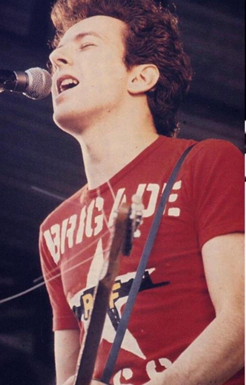 Pu&ograve; includere: Un uomo con i capelli corti e castani canta in un microfono mentre tiene una chitarra. Indossa una maglietta rossa con la scritta "BRIGADE" stampata in lettere bianche.