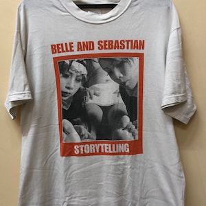 May include: White t-shirt featuring a black and white photograph of two people. The photo is within a red rectangle with the text "Belle and Sebastian" above the photo and "Storytelling" below the photo.