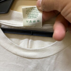 Puede incluir: Una camiseta blanca con un dise&ntilde;o de rayas verdes y blancas. La etiqueta de la camiseta dice "Este es un producto totalmente natural. No se han utilizado productos qu&iacute;micos en su fabricaci&oacute;n. T&uacute; tambi&eacute;n puedes seguir cuidando el medio ambiente utilizando detergentes y l&iacute;quidos de lavado biodegradables."