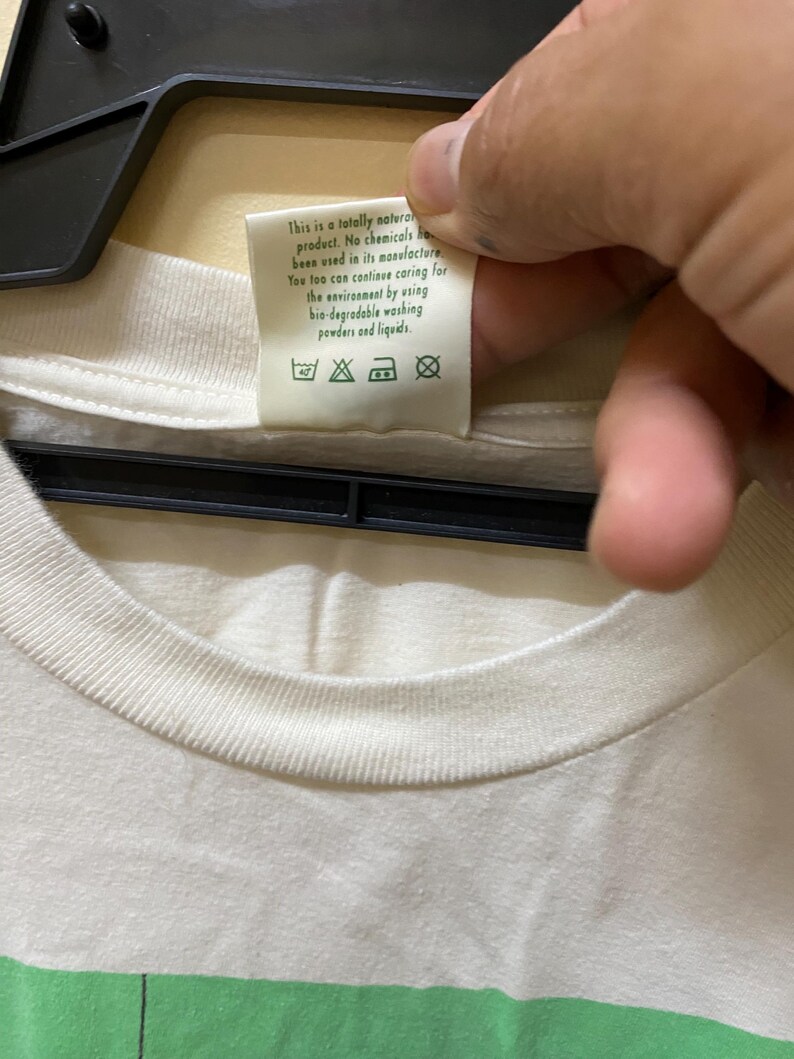 Puede incluir: Una camiseta blanca con un dise&ntilde;o de rayas verdes y blancas. La etiqueta de la camiseta dice "Este es un producto totalmente natural. No se han utilizado productos qu&iacute;micos en su fabricaci&oacute;n. T&uacute; tambi&eacute;n puedes seguir cuidando el medio ambiente utilizando detergentes y l&iacute;quidos de lavado biodegradables."