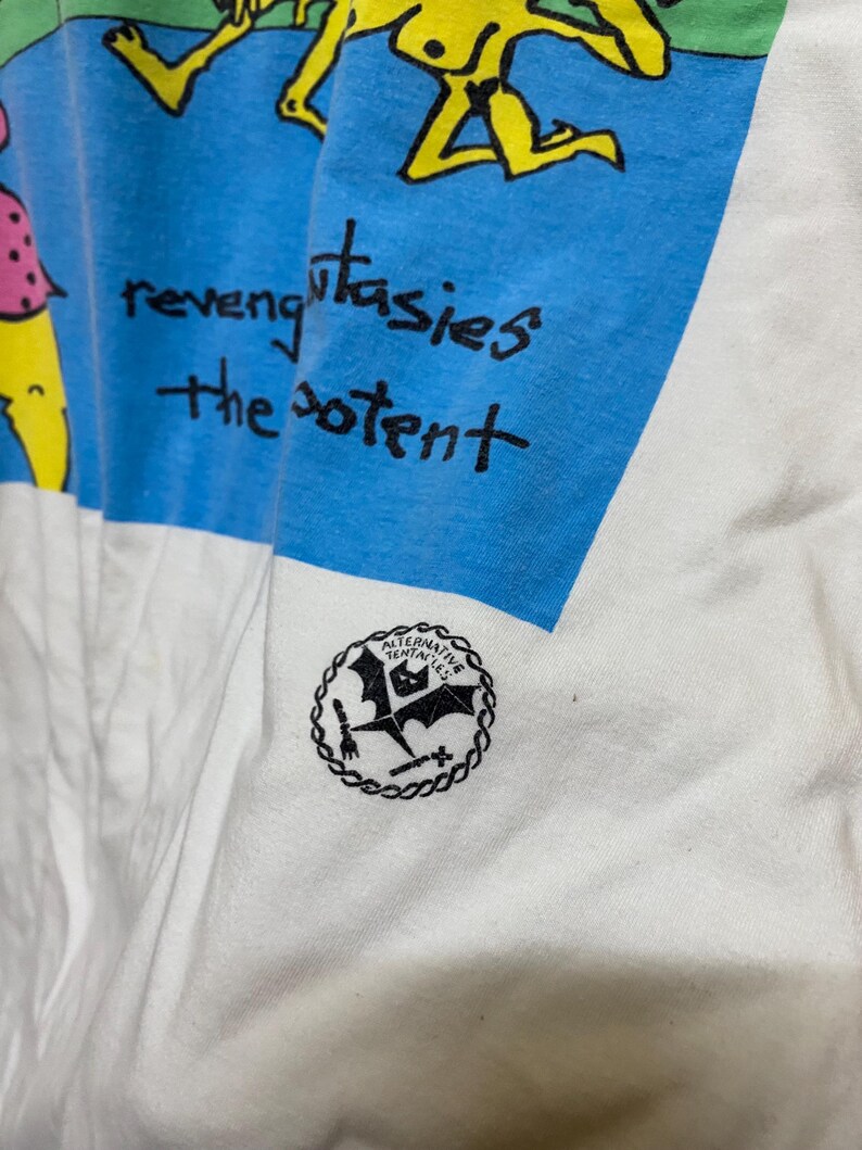 Puede incluir: Camiseta blanca con un gr&aacute;fico azul y verde de figuras de palitos. El texto "revenge fantasies the potent" est&aacute; escrito en negro. La parte inferior de la camiseta tiene un logotipo en blanco y negro que dice "Alternative Tentacles".