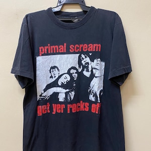 May include: Black t-shirt with a band graphic. The graphic features a black and white photo of a band with the text "primal scream get yer rocks off" in red.