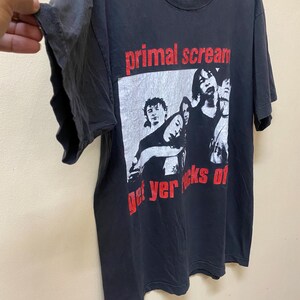 May include: Black t-shirt featuring a white and black image of a band. The band's name "primal scream" is printed in red above the image. The text "get yer rocks of" is printed in red below the image.