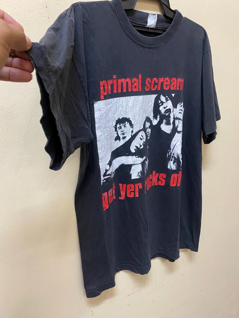 May include: Black t-shirt featuring a white and black image of a band. The band's name "primal scream" is printed in red above the image. The text "get yer rocks of" is printed in red below the image.