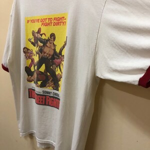 Puede incluir: Camiseta blanca con un ribete rojo y una impresi&oacute;n de p&oacute;ster de pel&iacute;cula vintage. El p&oacute;ster presenta a un hombre con una chaqueta de cuero y el texto "If you've got to fight - fight dirty!" y "The Street Fighter".