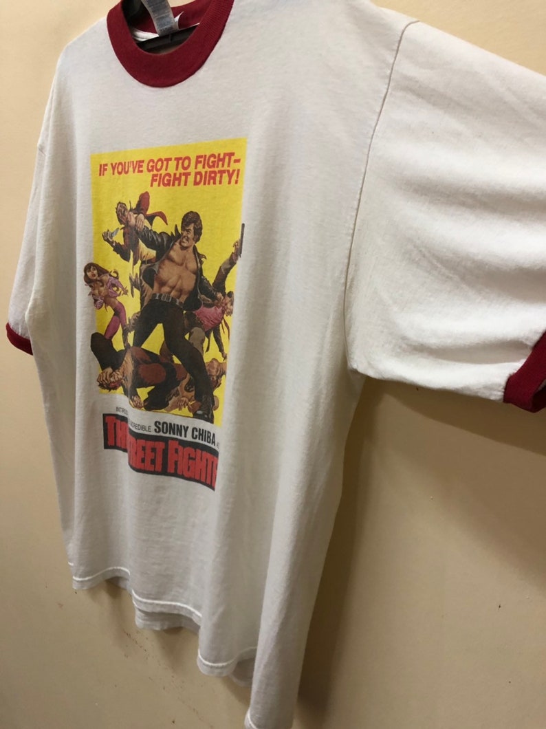 Puede incluir: Camiseta blanca con un ribete rojo y una impresi&oacute;n de p&oacute;ster de pel&iacute;cula vintage. El p&oacute;ster presenta a un hombre con una chaqueta de cuero y el texto "If you've got to fight - fight dirty!" y "The Street Fighter".