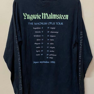 May include: Black long-sleeved t-shirt with a white print of Yngwie Malmsteen's name and the text "The Magnum Opus Tour". The tour dates and locations are listed below the text in white.