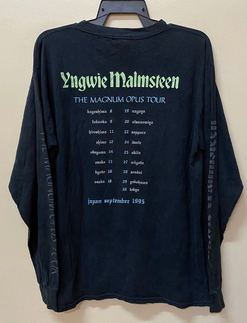 May include: Black long-sleeved t-shirt with a white print of Yngwie Malmsteen's name and the text "The Magnum Opus Tour". The tour dates and locations are listed below the text in white.