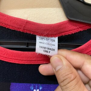 May include: A black and red t-shirt with a graphic print of a hand holding a glowing object. The tag says "100% Cotton, Made in Britain, Hand Wash Only."