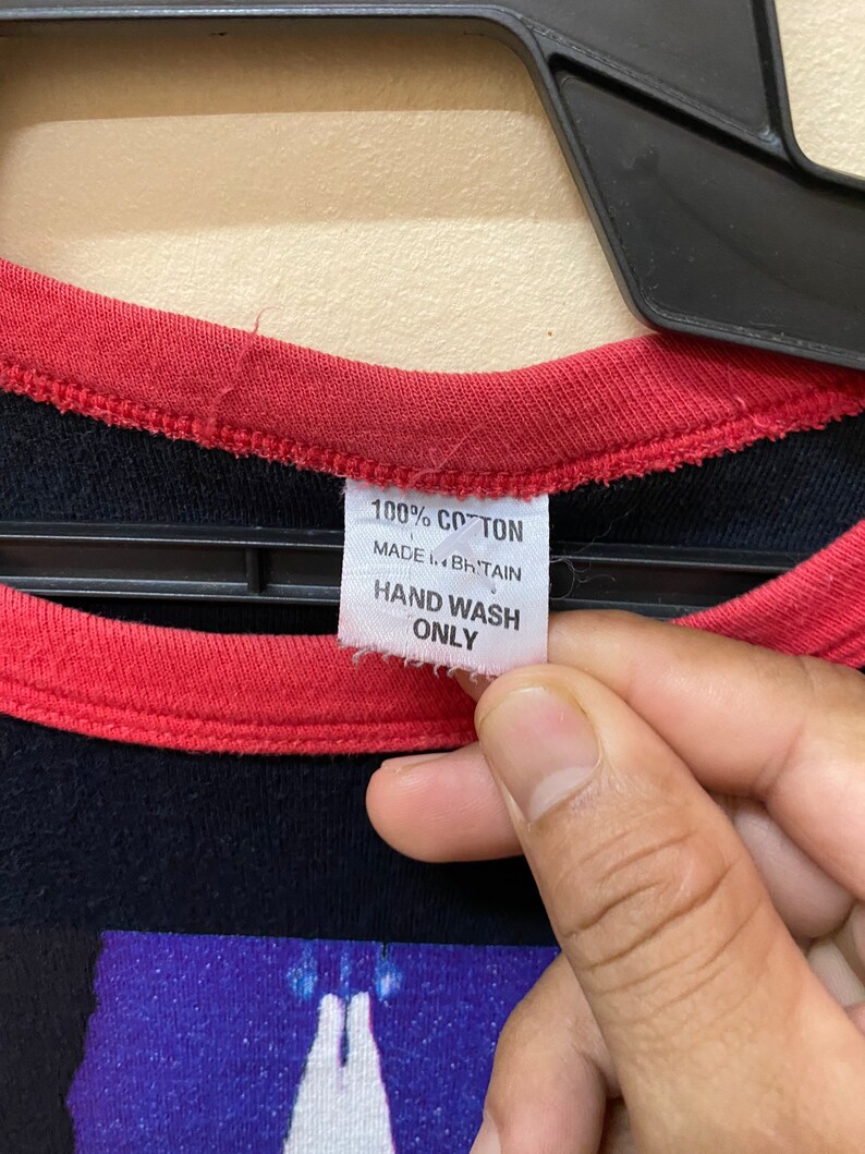 May include: A black and red t-shirt with a graphic print of a hand holding a glowing object. The tag says "100% Cotton, Made in Britain, Hand Wash Only."