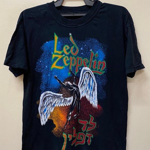 Op de afbeelding: Zwart T-shirt met een grafische print van een gevleugelde figuur en de tekst "Led Zeppelin" in groene en gele letters. De grafiek bevat ook Hebreeuwse tekst.