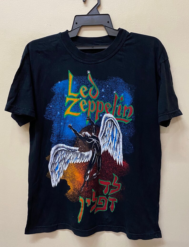 Op de afbeelding: Zwart T-shirt met een grafische print van een gevleugelde figuur en de tekst "Led Zeppelin" in groene en gele letters. De grafiek bevat ook Hebreeuwse tekst.