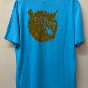 May include: A light blue t-shirt with a brown graphic of a cartoon face with a single eye and a tongue sticking out. The face is surrounded by a circle.