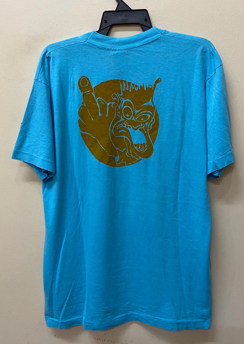 May include: A light blue t-shirt with a brown graphic of a cartoon face with a single eye and a tongue sticking out. The face is surrounded by a circle.