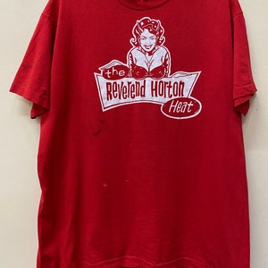 May include: Red t-shirt with a white graphic print of a woman with horns and a red dress. The graphic says "the Reverend Horton Heat".