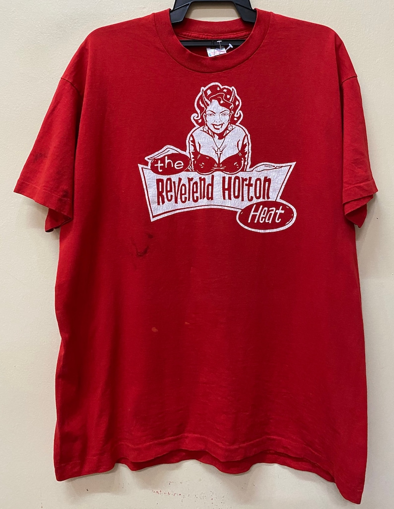 May include: Red t-shirt with a white graphic print of a woman with horns and a red dress. The graphic says "the Reverend Horton Heat".